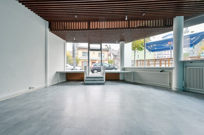 Empty room with tiled floor, white walls, wooden ceiling, glass doors, and large windows with a view of the street outside.