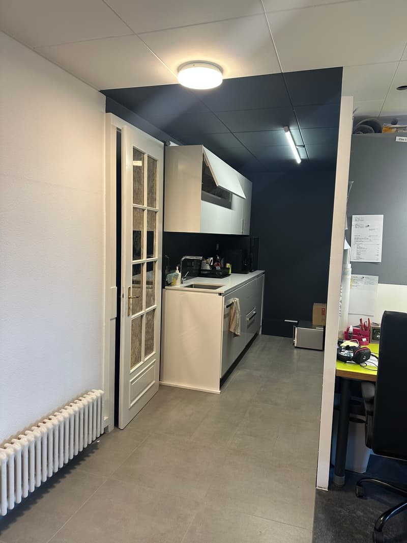 White cabinets, dark countertops, gas stove, white sink, overhead lighting, white tiled floor, open door to adjacent room with office desk, radiator, box on floor