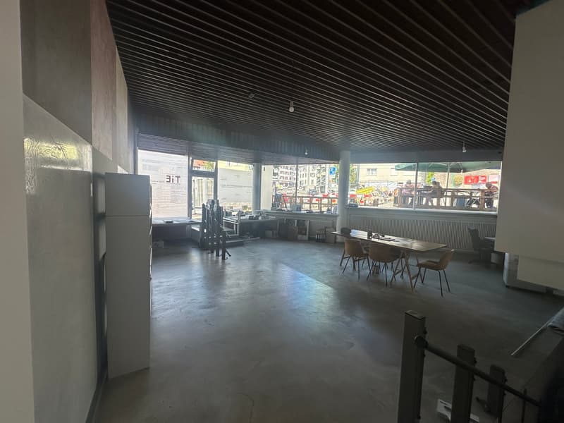 Empty large room, concrete floor, gray walls, staircase, ceiling with wooden beams, multiple windows, chairs, tables, white fridge, white railings.