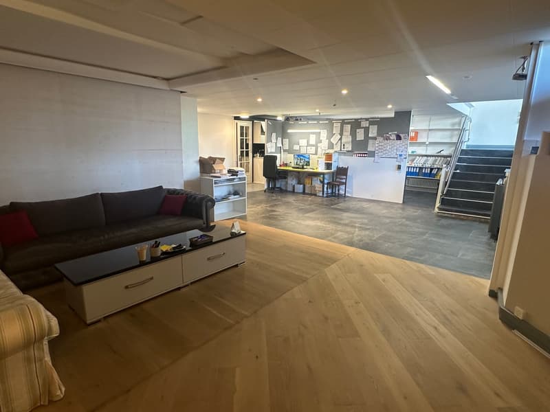 Spacious living room with hardwood floors, large brown sofa, black coffee table, white shelves, stairs leading to the upper floor, office area with desk and chair.