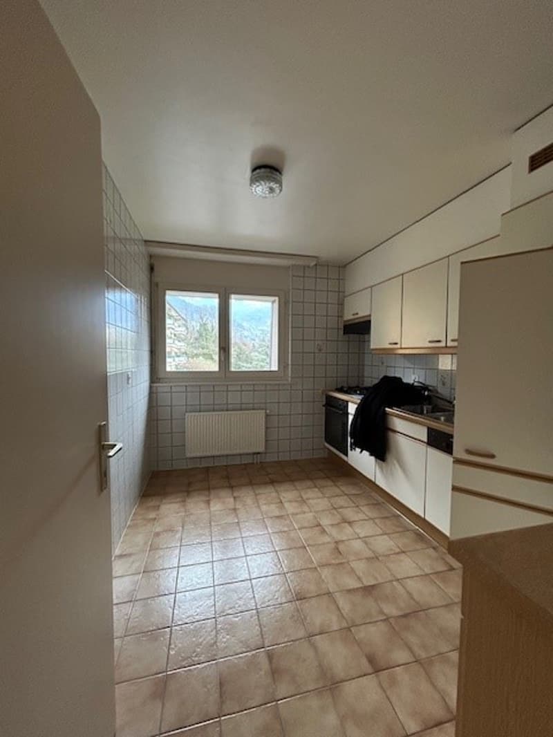An empty kitchen with white tiles, white cabinets, ceiling light, windows, stove, sink, and countertop.