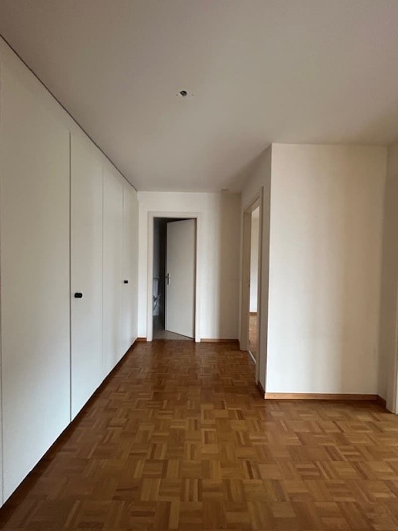 The image shows a long, narrow hallway with white walls and a wooden parquet floor. There are built-in closets or cabinets lining one side of the hallway, and a doorway leading to another room can be seen at the end of the hallway.
