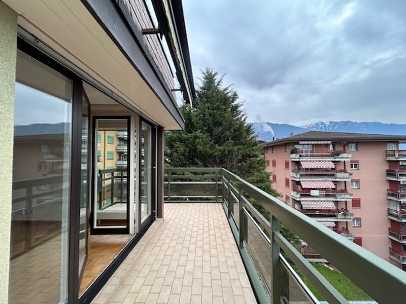 The image shows a balcony or terrace of an apartment building. The balcony has glass sliding doors and a railing. The building has a modern design with a mix of light and dark colors. There are other apartment buildings visible in the background, surround
