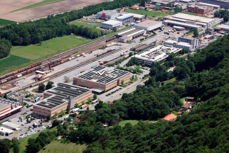An industrial building with solar panels, next to a railway line, surrounded by trees and other buildings