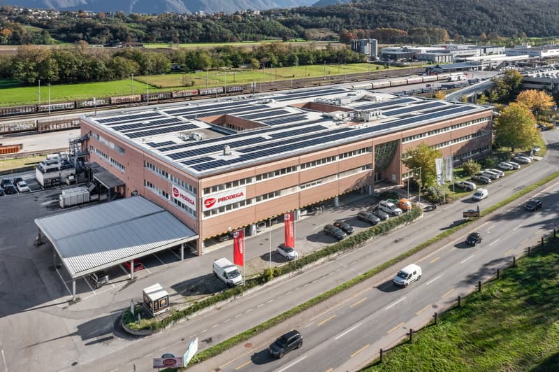 Solar panels on roof, several cars parked in front, expansive road, greenery, mountains in the distance.