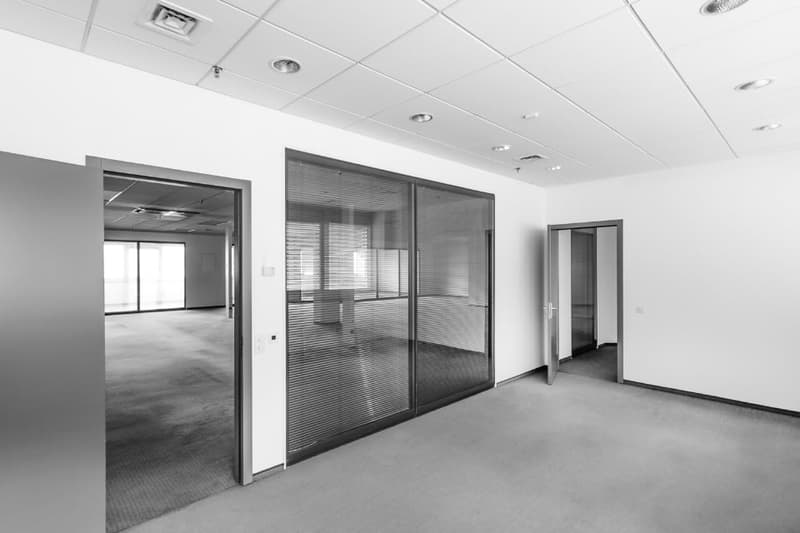 Empty office room with sliding glass doors, square tiles, white walls, two doors, carpeted floor, and overhead lights.