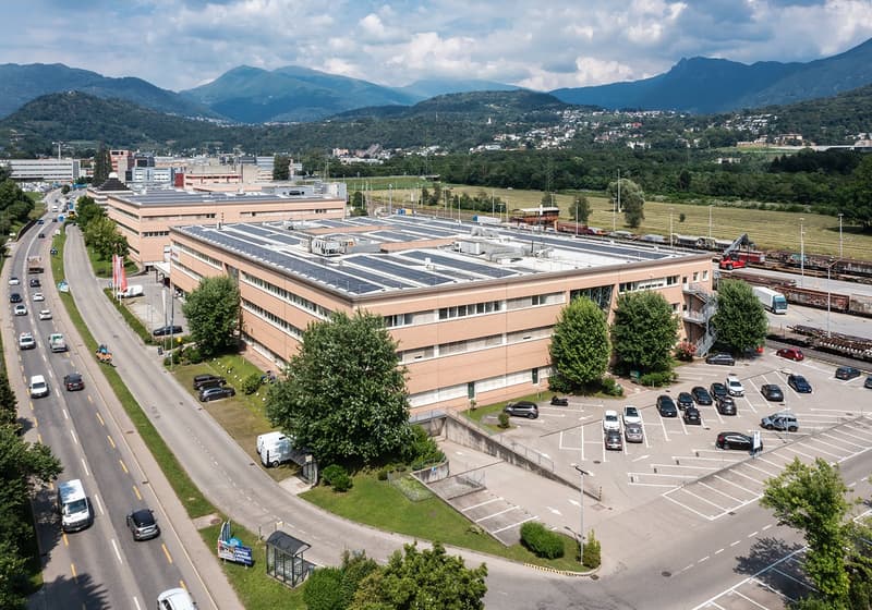 Industrial building with solar panels on the roof, several floors, large parking lot, cars driving on road, trees and plants surrounding the area