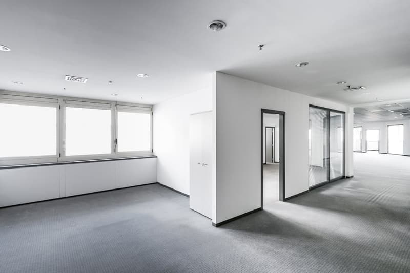an empty room with white walls and gray flooring, large glass windows, and several doors