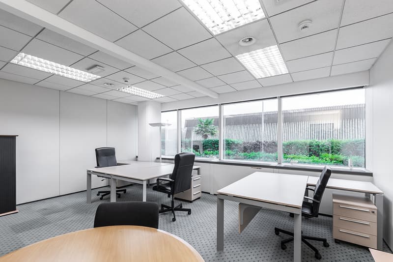 modern office room with white desks, black chairs, white walls, white ceiling with ceiling lights, large windows with view to outdoor