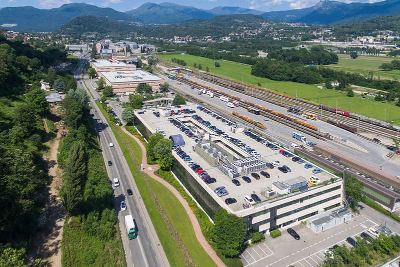 multilevel building with parking spaces, train tracks, road, trees, mountains in the background