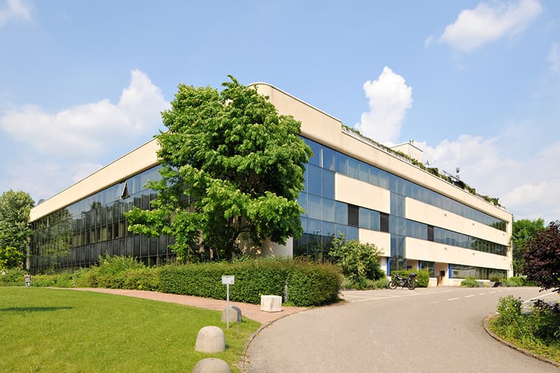 Modern building, large windows, glass facade, roof garden, front yard with trees, paved driveway, motorcycles parked.