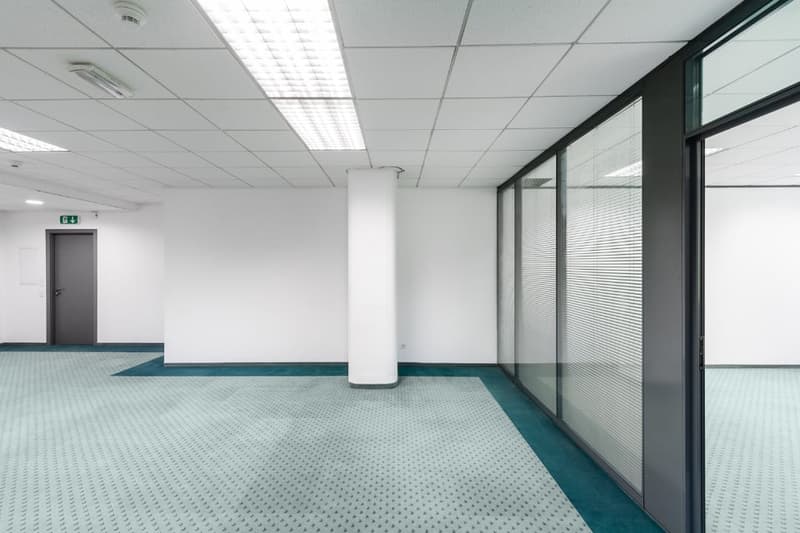 Empty room with ceiling lights, central column, gray door, glass windows, turquoise carpet