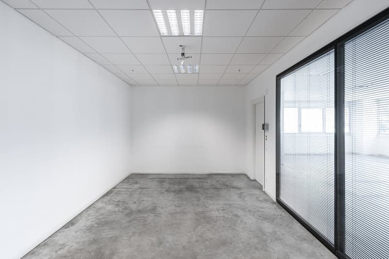 An empty room with a concrete floor and white walls, large window with blinds, open door
