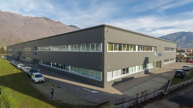 Industrial building with multiple windows, grey exterior, two levels, multiple cars parked in front, mountains in the background