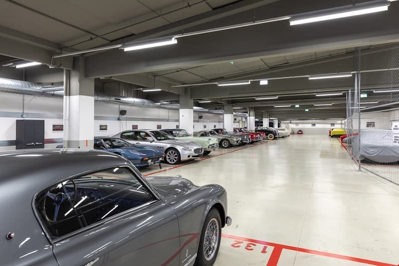 Underground parking garage, several vintage cars parked, white flooring, fluorescent lights, metal beams and pillars, security cameras, entrance/exit at the back