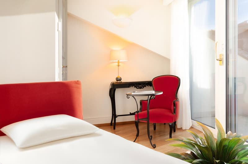 Single bed, white bedsheet, red headboard, red armchair, black desk with lamp, glass doors to balcony