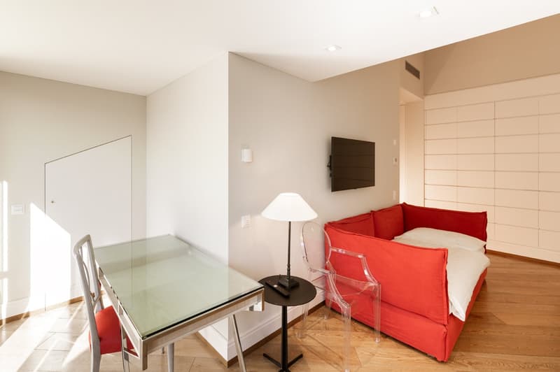 room with a glass table, a red sofa, a side table, a clear lamp, a remote control, two white chairs, a glass wall lamp, and a TV