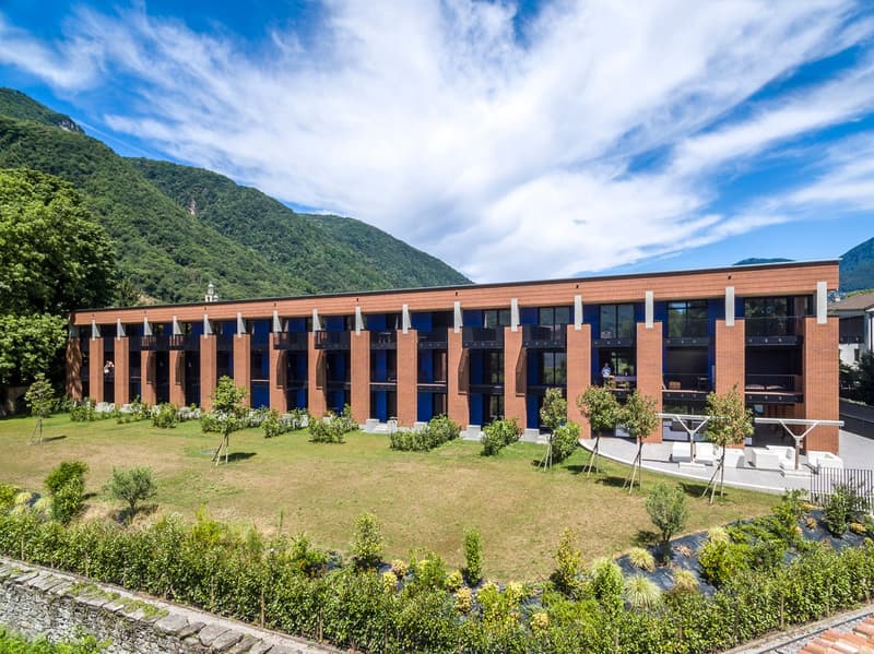 A multi-story brick building with balconies, surrounded by a well-maintained garden and landscaping. The building is situated in a scenic mountainous area with a blue sky and clouds overhead.
