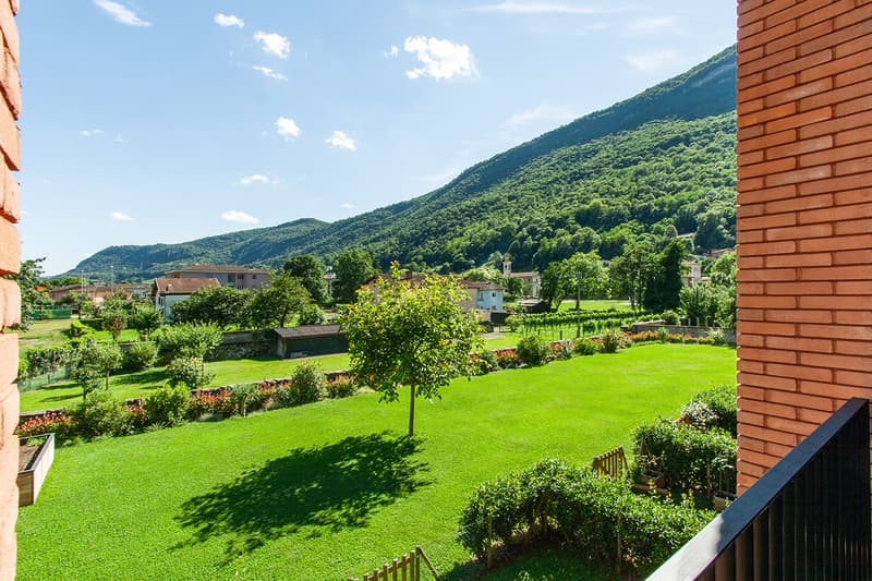 The image shows a scenic view of a lush, green landscape with rolling hills and mountains in the background. The foreground features a well-manicured garden with a grassy lawn, trees, and various shrubs and plants. The buildings in the distance suggest a