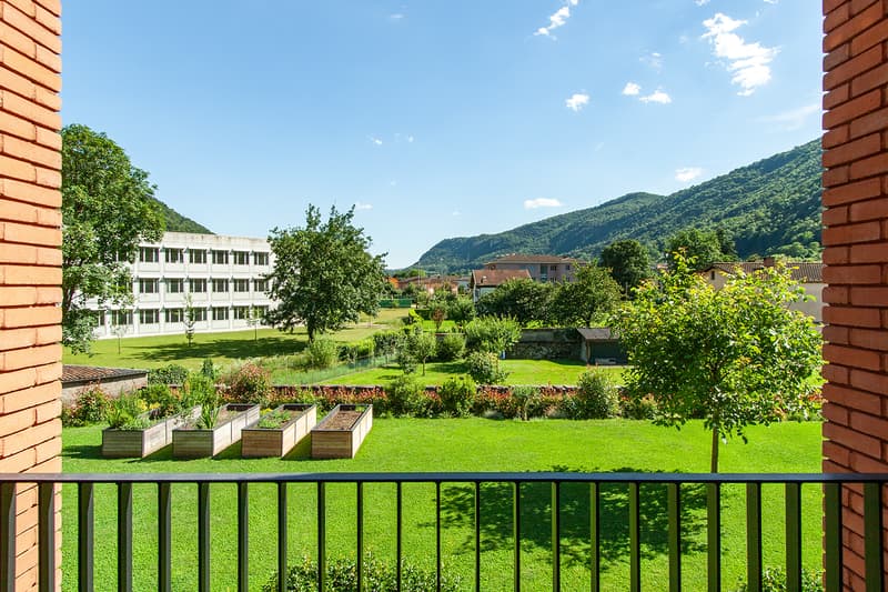 The image shows a multi-story residential building surrounded by a lush, green landscape. There is a balcony or terrace visible, and the building overlooks a scenic view of the mountains and trees in the distance.
