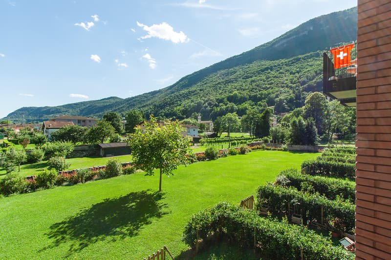 The image shows a scenic landscape with a lush, green garden and lawn surrounded by mountains covered in dense vegetation. There is a brick building with a Swiss flag visible, suggesting this is a location in Switzerland. The overall scene depicts a peace