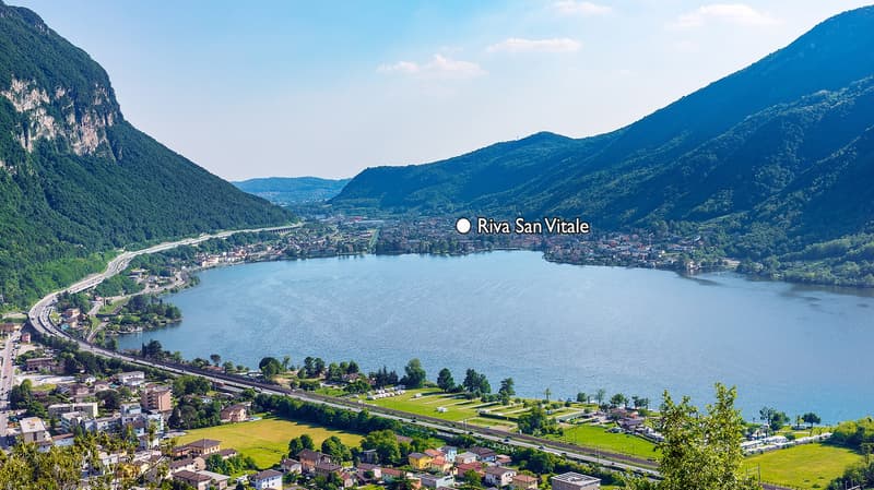 Aerial view of the town of Riva San Vitale situated on the shores of a large lake surrounded by mountains. The town appears to be nestled in a valley with a winding road passing through it. The landscape is lush and green, with a mix of residential and co