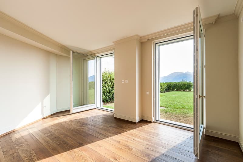 Spacious living room with large windows overlooking a grassy outdoor area, hardwood flooring, and neutral-colored walls