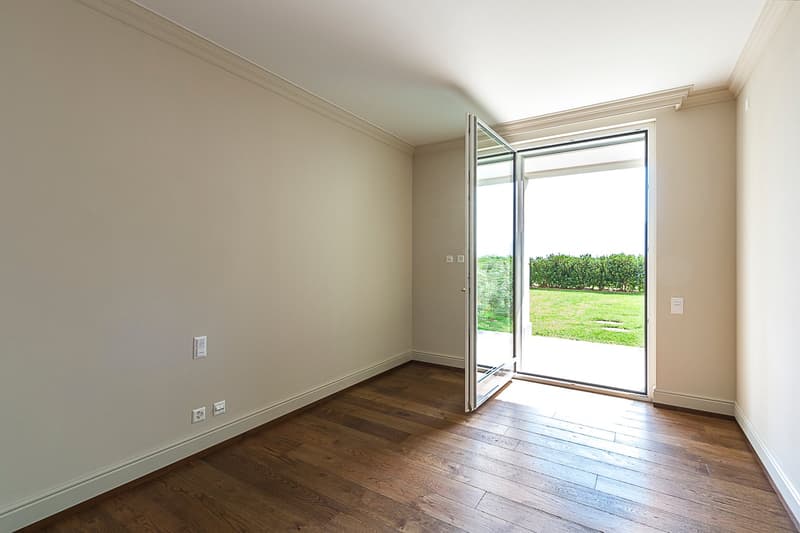 Spacious room with hardwood flooring, beige walls, and large sliding glass doors leading to a balcony or terrace overlooking a grassy outdoor area.