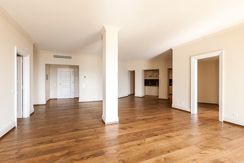 This image shows a spacious apartment interior with a large open floor plan. The room has hardwood floors, white walls, and a central column. There are multiple doorways leading to other rooms, including what appears to be a kitchen area. The overall desi