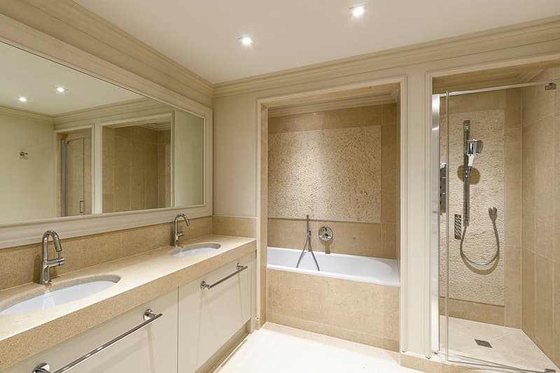Spacious bathroom with double vanity sinks, large mirror, bathtub, and separate shower area with glass enclosure. The bathroom features beige tile walls and floors, and built-in storage cabinets.