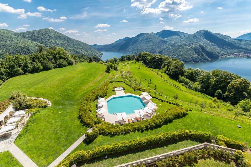 Aerial view of a luxury property with a large swimming pool surrounded by lush green lawns and gardens, overlooking a scenic lake and mountains in the background.