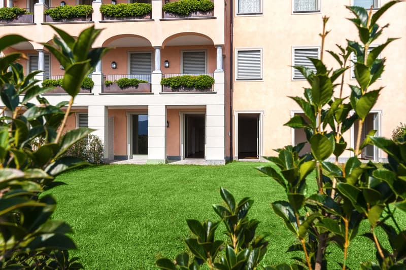3-story apartment building with arched windows, balconies with potted plants, and a well-manicured lawn in the foreground.