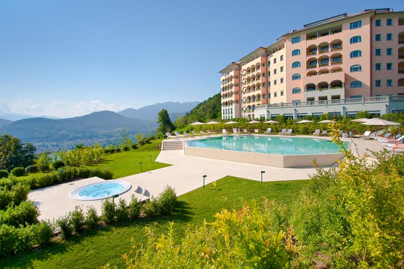 Multi-story hotel building with balconies, surrounded by lush green gardens and lawns. Large outdoor swimming pool with lounge chairs. Scenic mountain views in the background.