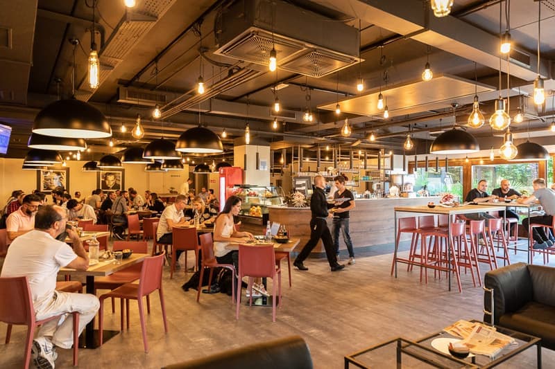 Spacious restaurant interior with high ceilings, exposed ductwork, and numerous hanging light fixtures. Tables and chairs are arranged throughout the dining area, and there is a bar and open kitchen visible. The restaurant appears to be busy with customer