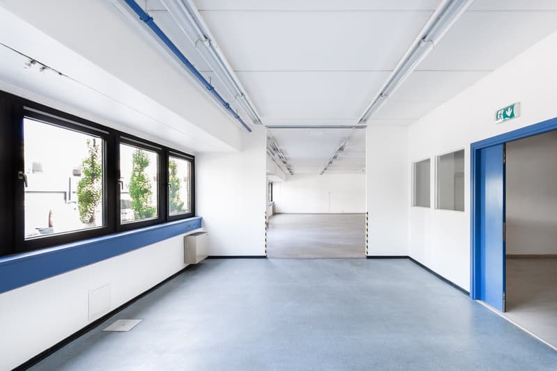 Large open office space with white walls, gray flooring, and large windows. The space has a clean, modern design with blue accents on the window frames and doors. The ceiling has exposed ductwork and lighting fixtures. There is an exit sign visible, indic