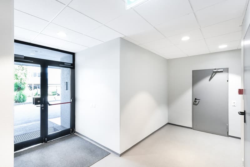 The image shows a modern, minimalist office space with a glass entrance door, white walls, and a gray tiled floor. The room has a clean and professional appearance, with a door leading to another area of the office.