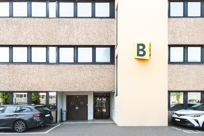 2-story office building with a granite-like exterior, large windows, and a prominent letter 'B' sign. There are two cars parked in the front of the building.