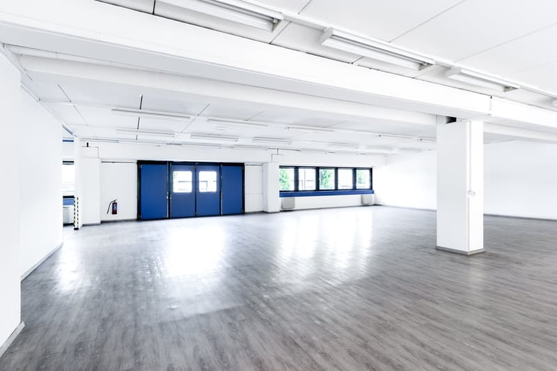 Large open commercial space with high ceilings, white walls, and gray hardwood flooring. There are blue garage doors at the far end of the room, indicating a garage or loading area.
