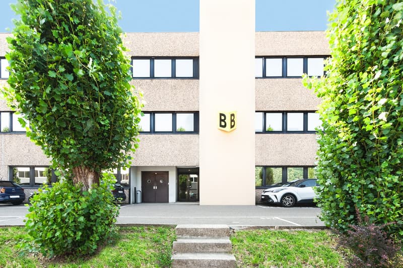 3-story office building with beige exterior, large windows, and a prominent 'BB' sign. The building is surrounded by lush greenery, including trees and shrubs. There is a parking area in front of the building.