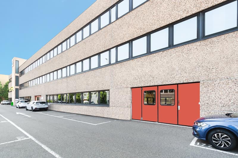 Multi-story commercial building with a textured concrete exterior, large windows, and red double doors. There are several parked cars in the parking lot in front of the building.