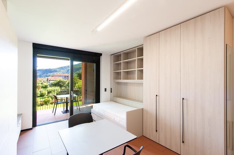 Spacious bedroom with large windows and sliding glass door leading to a balcony, providing a scenic view of the surrounding landscape. The room features built-in wooden cabinets and shelves, a bed, and a desk area.