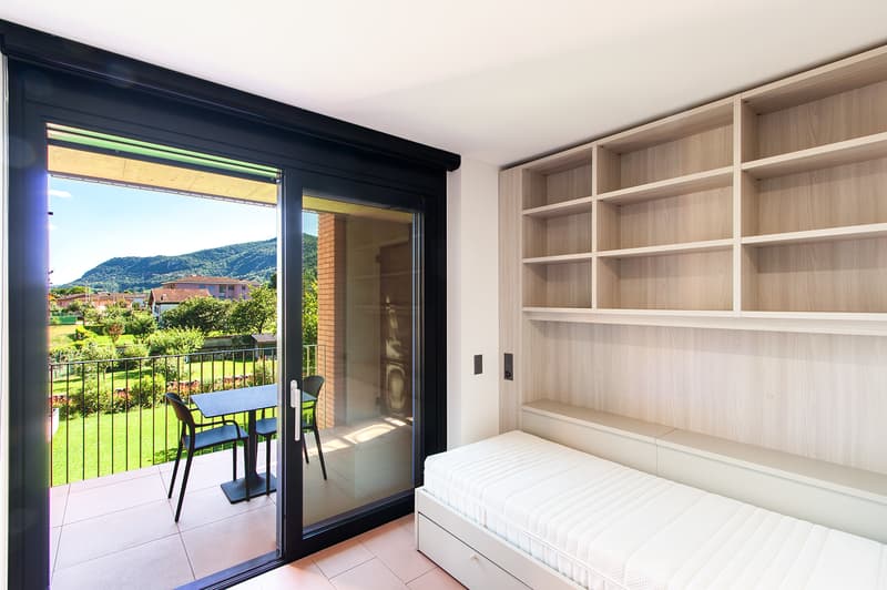 A modern room with sliding glass doors leading to a balcony, a table and chairs on the balcony, and a built-in bed against the wall