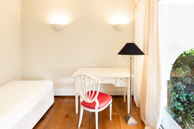 white walls, wooden desk, chair with red seat, white mattress, floor lamp
