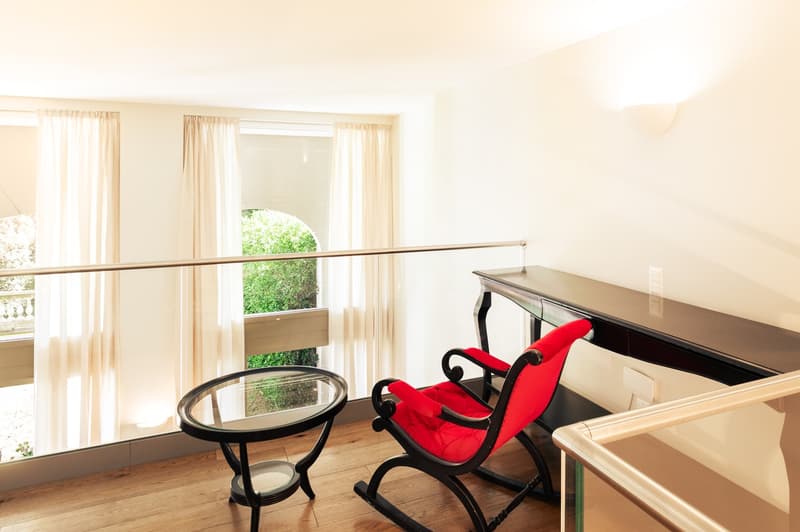 Balcony with a red chair, a glass table, a black desk with wall outlet, glass railings, and white curtains covering the window