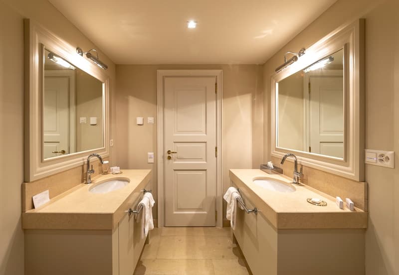 Modern bathroom with two vanities, mirrors with lights, two sinks, white cabinets, white towels on towel racks, floor outlet, open door, tiled floor.