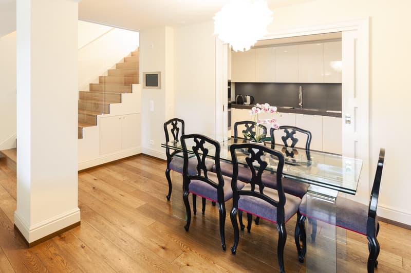 glass dining table, glass chairs, wooden floor, white walls, kitchen with white cabinets, stainless steel sink, staircase with glass railing
