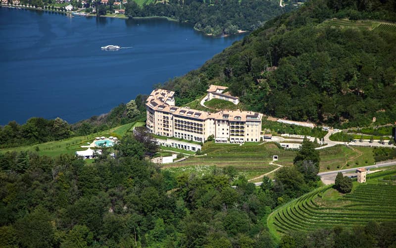 Hotel building on hill with view of lake, green surroundings, solar panels on roof, pool, terraced garden, boat on lake