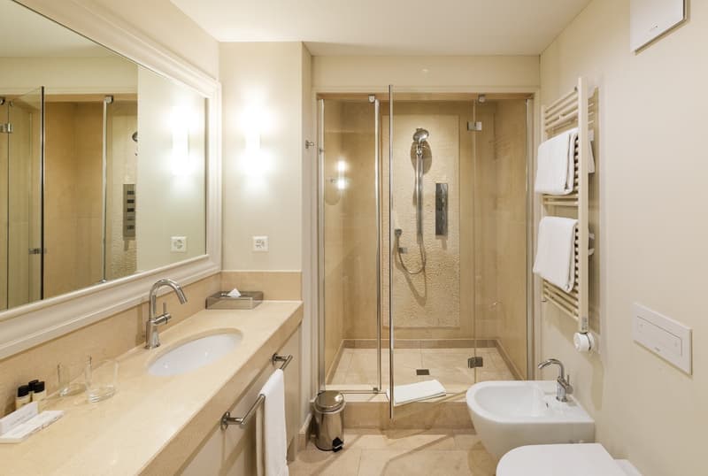 marble counter, glass walk-in shower, bidet, vanity with sink and mirror, toilet, towel racks, trash bin, glass cup holders, toiletries, power outlets
