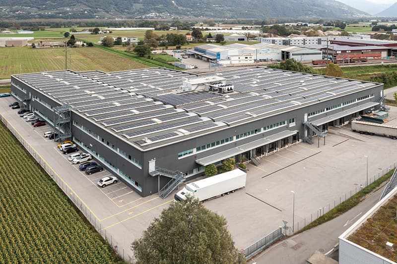 Large industrial warehouse or distribution center with a flat roof, multiple loading docks, and a parking lot in front of the building. The building is surrounded by agricultural fields and other industrial facilities in the background.