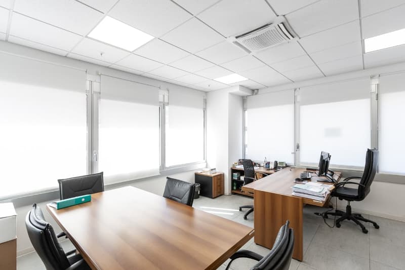 The image shows a modern, well-lit office space with a large conference table, several desks, and office chairs. The room has white walls, a tiled floor, and recessed lighting in the ceiling. There are large windows along one wall, providing natural light